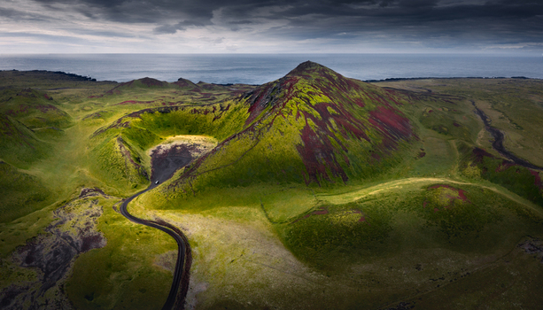 Aerial view of a vibrant volcanic crater and moss covered mountain peak with a winding road and the ocean under a dramatic sky in Iceland.