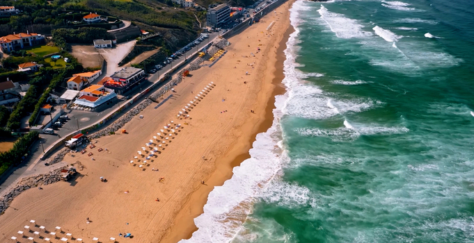 Aerial view of a sandy beach with white umbrellas turquoise waves crashing on the shore and coastal buildings along the road in Sintra Lisboa Portugal. Aerial view of a sandy beach with white umbrellas turquoise waves crashing on the shore and coastal buildings along the road in Sintra Lisboa Portugal.