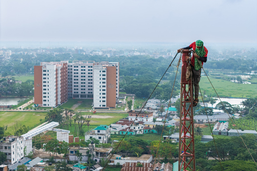 Trishal Upazila Bangladesh 13 May 2026 Aerial view of a worker climbing a tall metal tower with a large modern building and lush green landscape in the background. Trishal Upazila Bangladesh 13 May 2026 Aerial view of a worker climbing a tall metal tower with a large modern building and lush green landscape in the background.