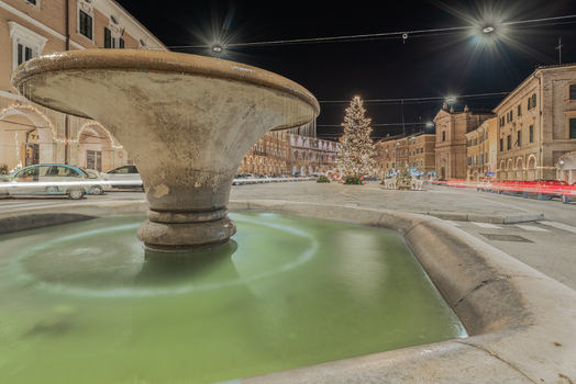 View of a stone fountain in the town square at night with an illuminated Christmas tree historic buildings and light trails from passing cars in San Severino Marche Marche Italy. View of a stone fountain in the town square at night with an illuminated Christmas tree historic buildings and light trails from passing cars in San Severino Marche Marche Italy.