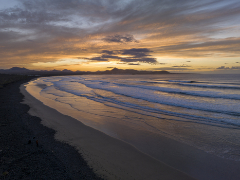 Aerial view of a beach with dark volcanic sand and gentle waves at sunset with mountains in the background under a dramatic golden sky in Lanzarote Canarias Spain. Aerial view of a beach with dark volcanic sand and gentle waves at sunset with mountains in the background under a dramatic golden sky in Lanzarote Canarias Spain.