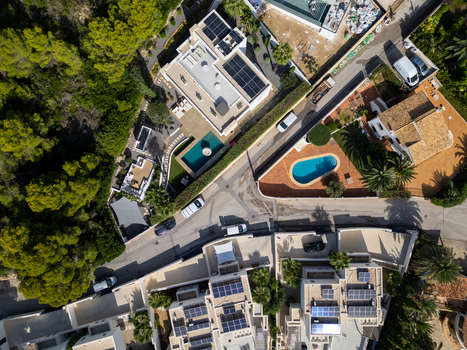 Aerial view of residential villas with swimming pools and solar panels on rooftops surrounded by lush green trees and roads in Moraira Valencian Community Spain.