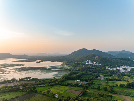 Aerial view of the serene lake surrounded by lush green fields rolling hills and scattered buildings under a soft sunset sky in Udaipur Rajasthan India.