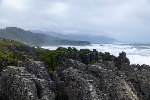 View of rugged layered pancake rocks meet the turbulent foamy surf under a grey brooding sky Pancake Rocks and Blowholes Walk West Coast Region New Zealand. View of rugged layered pancake rocks meet the turbulent foamy surf under a grey brooding sky Pancake Rocks and Blowholes Walk West Coast Region New Zealand.