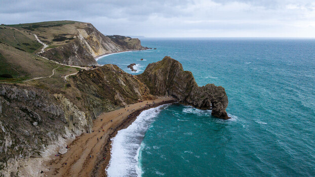 Aerial view of the iconic Durdle Door natural limestone arch on the Jurassic Coast with people on the beach and cliffside paths under a cloudy sky in Wareham England United Kingdom. Aerial view of the iconic Durdle Door natural limestone arch on the Jurassic Coast with people on the beach and cliffside paths under a cloudy sky in Wareham England United Kingdom.