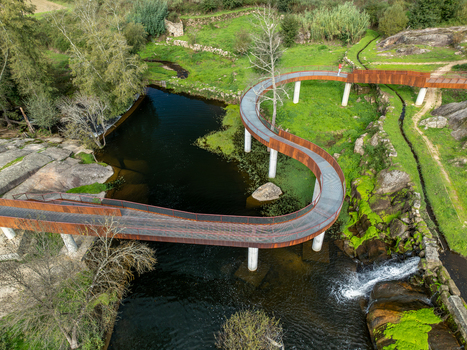Aerial view of the Acude da Ribeira curved walkway with a person walking across the river surrounded by lush green vegetation and rocky terrain in Ervedal Coimbra Portugal. Aerial view of the Acude da Ribeira curved walkway with a person walking across the river surrounded by lush green vegetation and rocky terrain in Ervedal Coimbra Portugal.