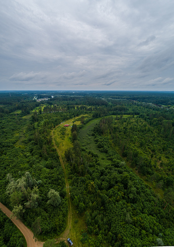 Aerial view of a dense green forest landscape featuring a winding dirt road and a small red structure under a vast cloudy sky in Latvia. Aerial view of a dense green forest landscape featuring a winding dirt road and a small red structure under a vast cloudy sky in Latvia.