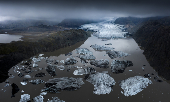 Aerial view of a massive glacier tongue flowing into a lagoon filled with floating icebergs surrounded by dark volcanic mountains under a moody sky Iceland.