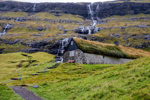 View of a traditional stone house with a grass roof nestled at the base of a steep cliff with cascading waterfalls in the Faroe Islands.