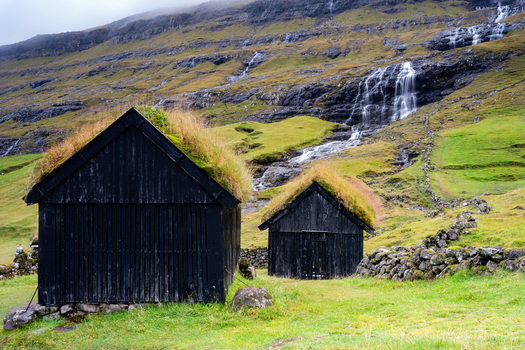 View of traditional black wooden sheds with turf roofs set against a grassy hillside with a waterfall in the Faroe Islands.