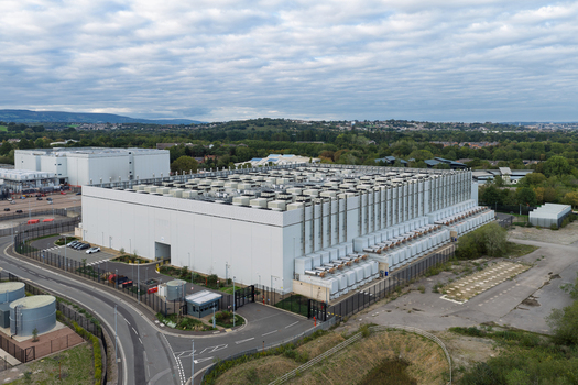 Aerial view of a large data center facility with extensive rooftop HVAC units surrounded by a secure fenced perimeter and access roads in the United Kingdom.