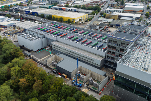 Aerial view of a large data center facility with colorful rooftop cooling units adjacent construction site and surrounding industrial area in the United Kingdom.