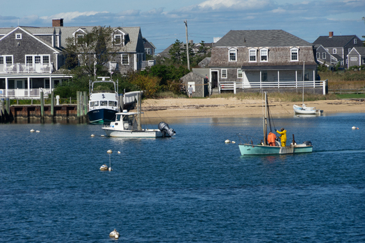 View of fishing boats in the harbor with fishermen in yellow and orange gear shingled houses and calm water on Nantucket Island in Nantucket Massachusetts United States.