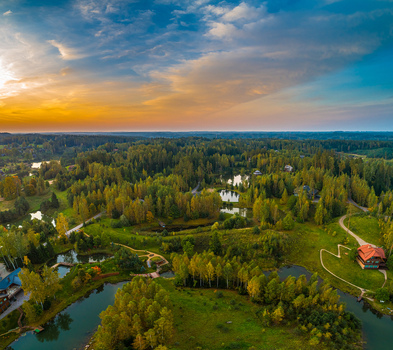 Aerial view of a lush green landscape featuring small ponds dense forests a house with a red roof and a vibrant sunset sky in Latvia. Aerial view of a lush green landscape featuring small ponds dense forests a house with a red roof and a vibrant sunset sky in Latvia.