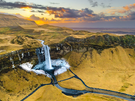 Aerial view of cascading waterfall plunges into icy pool amidst rugged landscape golden fields contrasting with dark cliffs Seljalandsfoss Rang&aacute;r&thorn;ing eystra Iceland.