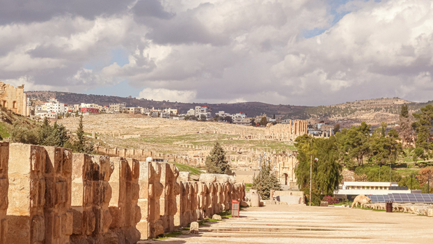 View of the ancient Roman ruins of Jerash with stone columns historic walls and the modern city in the background under a cloudy sky in Jerash Jerash Governorate Jordan. View of the ancient Roman ruins of Jerash with stone columns historic walls and the modern city in the background under a cloudy sky in Jerash Jerash Governorate Jordan.