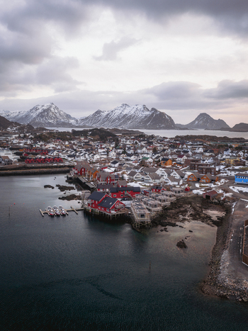 Aerial view of the fishing village nestling against snow dusted mountains the red roofed buildings contrasting with the dark water Ballstad Nordland Norway. Aerial view of the fishing village nestling against snow dusted mountains the red roofed buildings contrasting with the dark water Ballstad Nordland Norway.