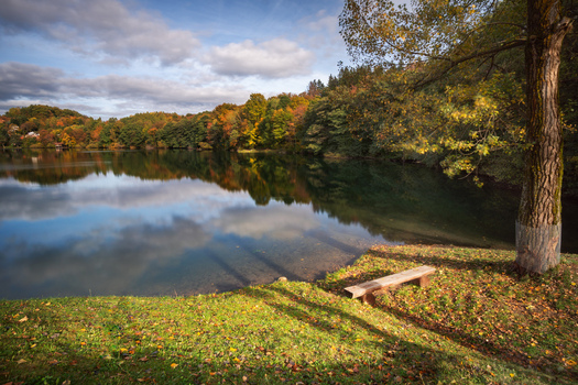 View of Velka Richnava Lake surrounded by colorful autumn trees and reflections in the water with a wooden bench in the foreground in Stiavnicke Bane Banska Bystrica Region Slovakia.