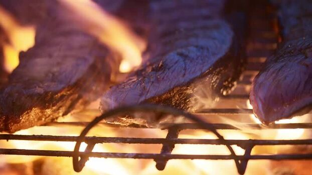 View of steaks grilling over open flames on a barbecue.