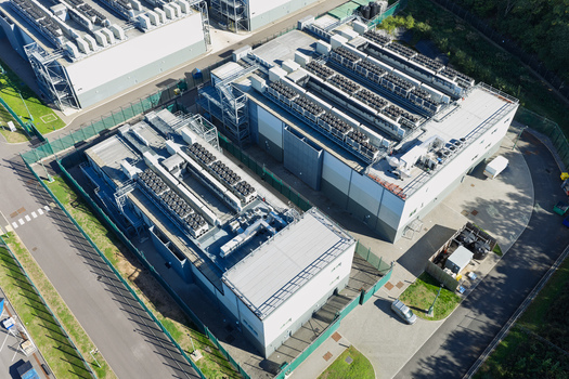 Aerial view of a modern data center facility with large cooling units on the rooftops surrounded by security fencing and paved access roads in the United Kingdom.
