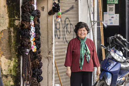 Hanoi Ha Noi Vietnam 11 November 2025 View of an elderly woman smiling in front of a shop displaying hair extensions with a blue motorbike parked nearby.
