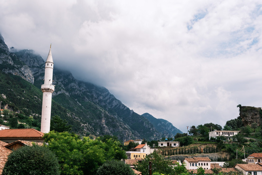 View of a white mosque minaret rising above red tiled rooftops and lush greenery with rugged mountains under a cloudy sky in Kruje Durres County Albania.