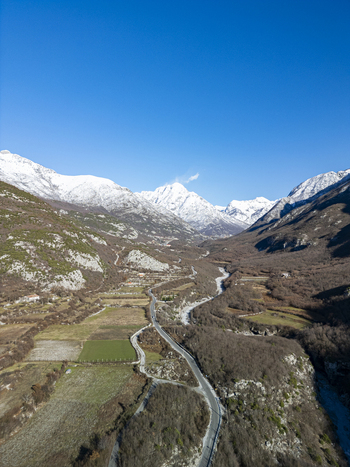 Aerial view of a winding road traversing a snow capped mountain valley with agricultural fields and rocky terrain in Bzhete Markaj Qarku i Shkodres Albania. Aerial view of a winding road traversing a snow capped mountain valley with agricultural fields and rocky terrain in Bzhete Markaj Qarku i Shkodres Albania.