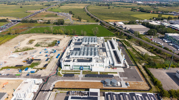 Aerial view of a modern hyperscale data center facility with cooling units and solar panels surrounded by industrial infrastructure and green fields in Melegnano Lombardy Italy.