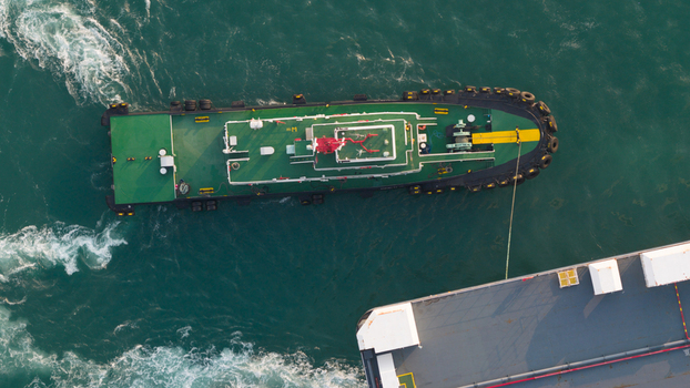 Aerial view of a green tugboat maneuvering alongside a large vessel in the turquoise waters of the industrial port in Laem Chabang Chon Buri Thailand.
