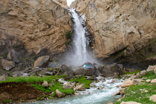 View of Khamosh Waterfall cascading down steep rocky cliffs into a mountain stream with green grass and boulders in Skardu Gilgit Pakistan.