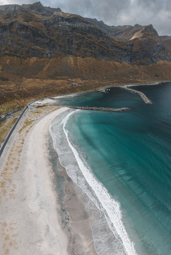 Aerial view of the sandy beach and turquoise waters of the coast with rugged mountains and breakwaters in Senja Troms Norway.