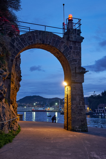 Pasai Donibane Spain 09 March 2026 View of the historic stone archway along the harbor street at dusk with a person sitting by the water and a red beacon light.