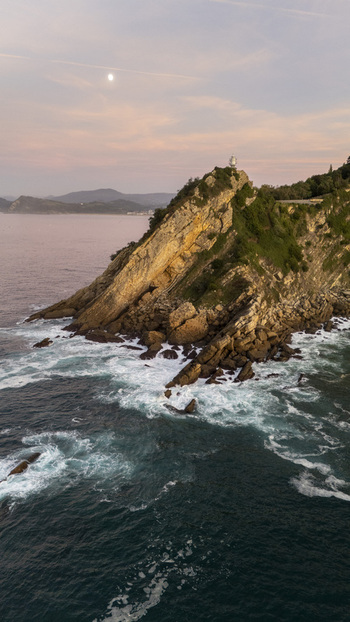 Aerial view of Getariako Itsasargia lighthouse perched on a rocky headland with crashing waves and the moon in the sky at sunset in Getaria Euskadi Spain. Aerial view of Getariako Itsasargia lighthouse perched on a rocky headland with crashing waves and the moon in the sky at sunset in Getaria Euskadi Spain.