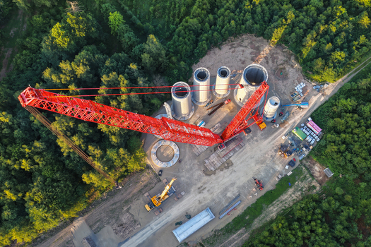 Aerial view of a large red crawler crane assembling wind turbine tower segments at a construction site surrounded by dense forest in Hofgeismar Hessen Germany. Aerial view of a large red crawler crane assembling wind turbine tower segments at a construction site surrounded by dense forest in Hofgeismar Hessen Germany.
