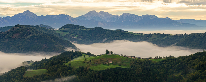 Aerial view of the Church of St. Thomas perched on a green hill above a sea of morning fog with the Julian Alps in the background in Selca Skofja Loka Slovenia. Aerial view of the Church of St. Thomas perched on a green hill above a sea of morning fog with the Julian Alps in the background in Selca Skofja Loka Slovenia.