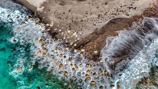 Aerial view of Tapuae Beach featuring dark sand scattered rocks and turquoise waves crashing against the shoreline in Omata Taranaki Region New Zealand.