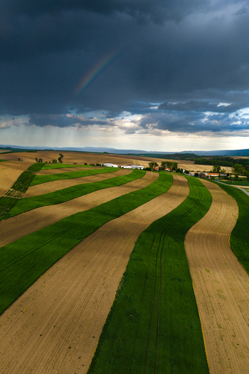 Aerial view of rolling agricultural fields with alternating green and brown strips under a dramatic stormy sky with a faint rainbow near Nasedlovice South Moravian Region Czechia.