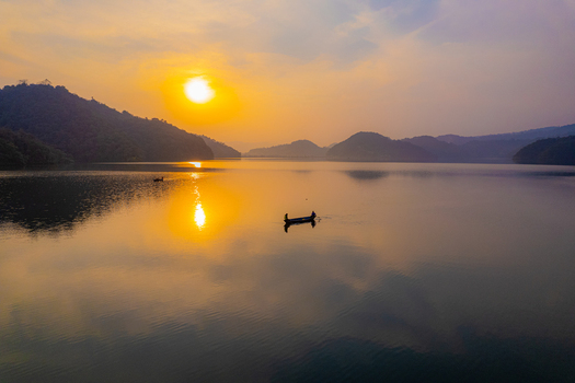 Aerial view of a small boat with people on a calm lake during sunset surrounded by rolling hills in Nepal.