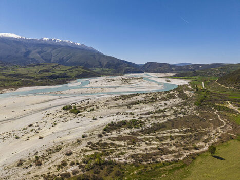 Aerial view of the Vjosa Wild River National Park with its wide braided riverbed lush green hills and snow capped mountains in the background in Permet Gjirokaster County Albania. Aerial view of the Vjosa Wild River National Park with its wide braided riverbed lush green hills and snow capped mountains in the background in Permet Gjirokaster County Albania.
