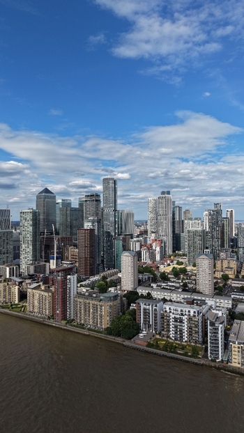 London United Kingdom 16 June 2026 Aerial view of Canary Wharf financial district featuring modern skyscrapers and residential blocks along the River Thames under a cloudy blue sky.