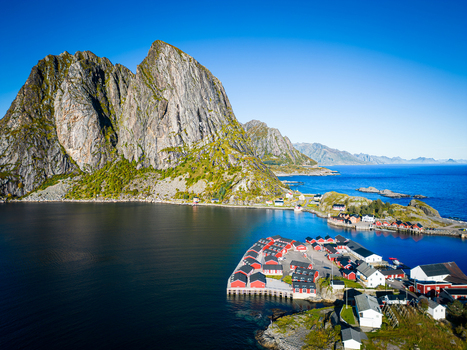 Aerial view of the traditional red rorbuer cabins on the waterfront surrounded by dramatic steep mountains and deep blue sea under a clear sky in Hamnoy Nordland Norway. Aerial view of the traditional red rorbuer cabins on the waterfront surrounded by dramatic steep mountains and deep blue sea under a clear sky in Hamnoy Nordland Norway.