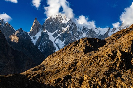 View of the snow capped peaks of the Hunza Valley illuminated by golden hour light with rugged mountain slopes and a deep blue sky in Karimabad GB Pakistan. View of the snow capped peaks of the Hunza Valley illuminated by golden hour light with rugged mountain slopes and a deep blue sky in Karimabad GB Pakistan.