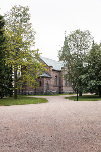 View of the red brick Jyvaskyla City Church with its tall spire surrounded by lush green trees and a gravel path in Jyvaskyla Central Finland Finland.