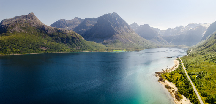 Aerial view of Kvaloya island featuring dramatic mountains deep blue fjord waters and a coastal road winding along the green shoreline in Tromso Municipality Troms Norway. Aerial view of Kvaloya island featuring dramatic mountains deep blue fjord waters and a coastal road winding along the green shoreline in Tromso Municipality Troms Norway.