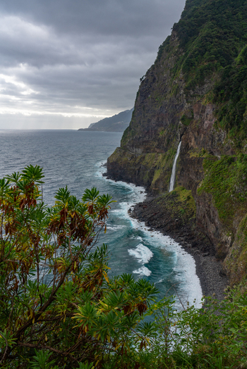 Aerial view of a waterfall cascading down a steep lush green cliff into the Atlantic Ocean with rocky coastline and cloudy sky in Madeira. Aerial view of a waterfall cascading down a steep lush green cliff into the Atlantic Ocean with rocky coastline and cloudy sky in Madeira.
