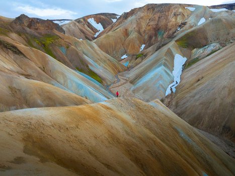 Aerial view of a solitary figure in red amidst the stark multi hued ridges and snow dusted peaks of Iceland s highlands a tapestry of ochre blue and cream Landmannalaugar Iceland.