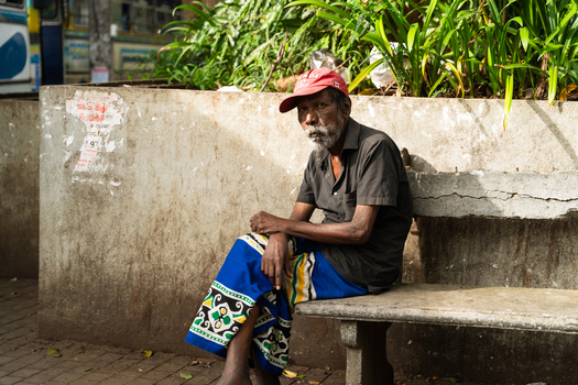 Sri Lanka 20 February 2026 View of an elderly man with a grey beard and red cap sitting on a stone bench wearing a traditional blue patterned sarong in an urban setting.