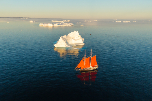 Aerial view of a sailboat with vibrant orange sails navigating through calm Arctic waters surrounded by majestic icebergs under the soft glow of the golden hour in Greenland. Aerial view of a sailboat with vibrant orange sails navigating through calm Arctic waters surrounded by majestic icebergs under the soft glow of the golden hour in Greenland.