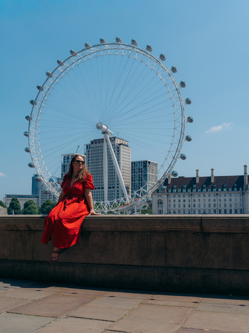 View of a woman in a vibrant red dress on a stone wall against the majestic London Eye under a clear blue sky London England United Kingdom. View of a woman in a vibrant red dress on a stone wall against the majestic London Eye under a clear blue sky London England United Kingdom.