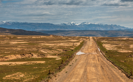 View of a dirt road stretching through the vast arid landscape with snow capped mountains in the distance under a cloudy sky in At Bashi District Naryn Region Kyrgyzstan. View of a dirt road stretching through the vast arid landscape with snow capped mountains in the distance under a cloudy sky in At Bashi District Naryn Region Kyrgyzstan.
